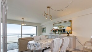 A dining area with a table set for four, a sofa, a large window with a view, and a chandelier. - Apartment in Blankenberge With Sea Views (Blankenberge)