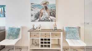 A room with a wooden desk, two white chairs, a painting of a woman by the sea, and a radiator. - Apartment in Blankenberge With Sea Views (Blankenberge)
