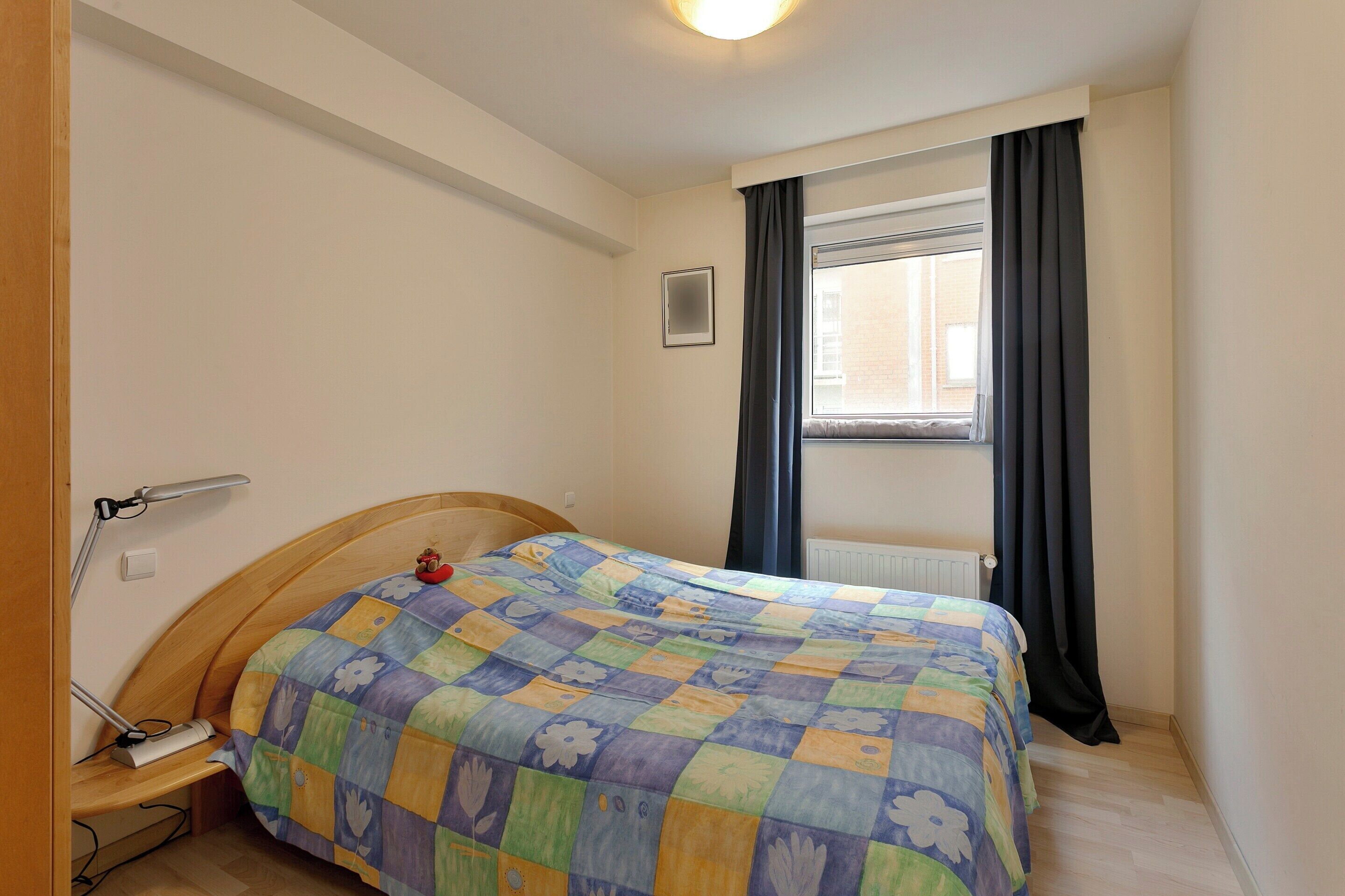 Un dormitorio pequeño y ordenado, con cama, mesita de noche, lámpara y ventana con cortinas.