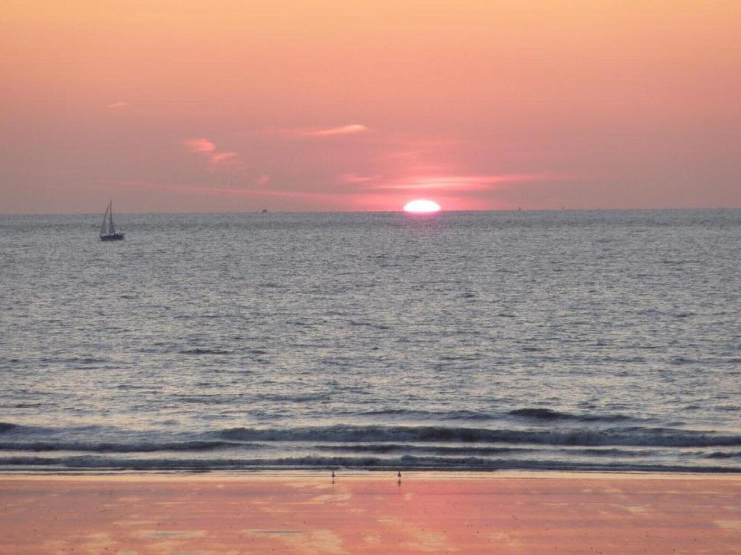 A sunset over a calm sea with a sailboat and a distant shoreline.