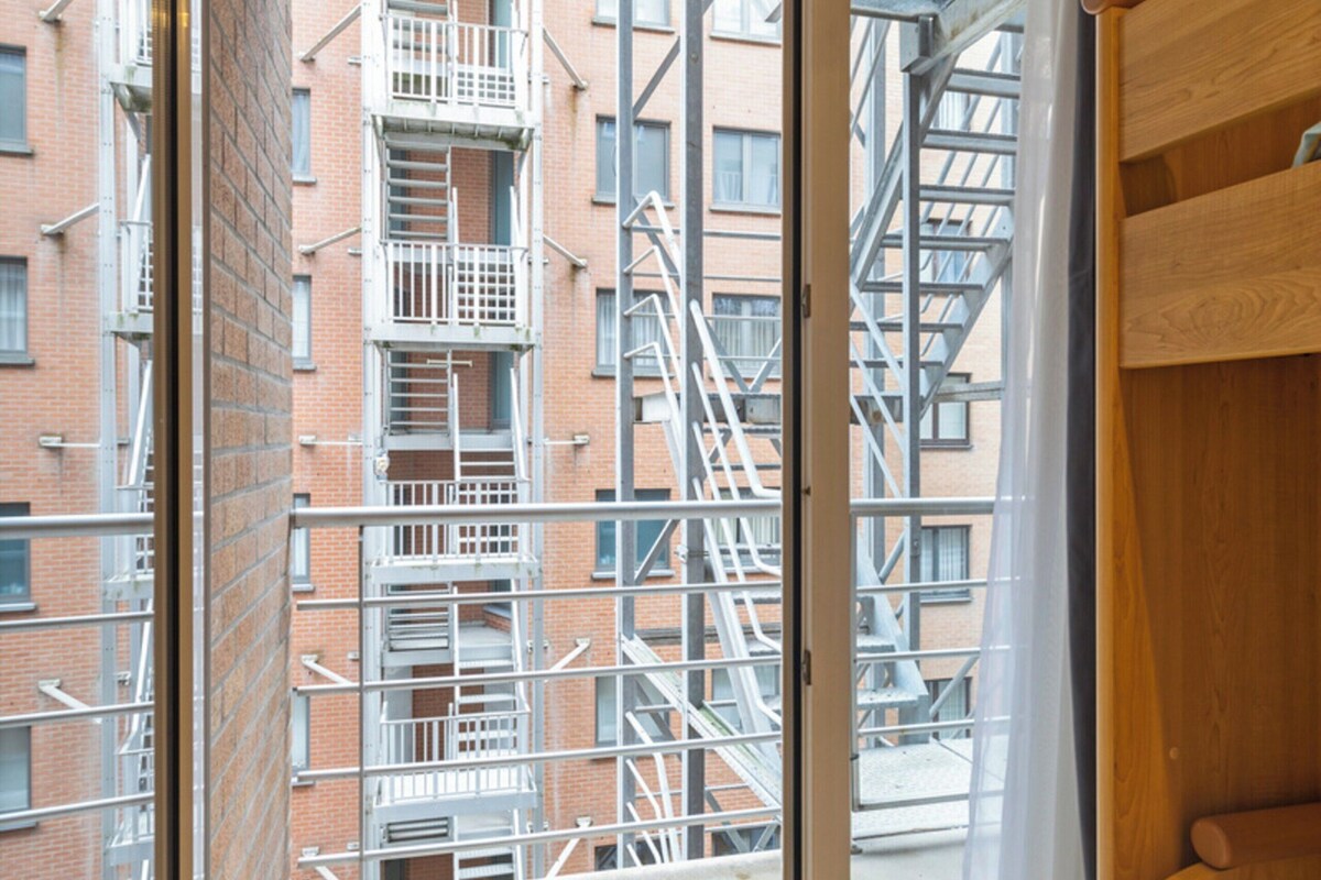 A room with a wooden bunk bed, a sliding glass door leading to a balcony with a view of a brick building and fire escapes.