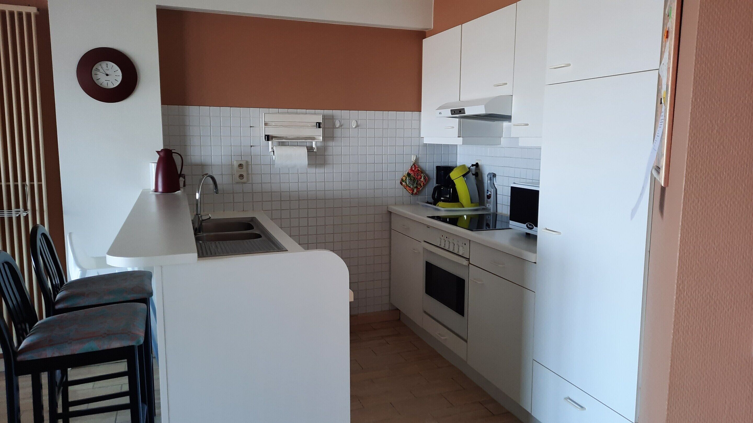 Une cuisine de taille réduite avec des meubles blancs, un four encastré, un four à micro-ondes et un évier. Il y a une horloge au mur et une chaise dans le coin.