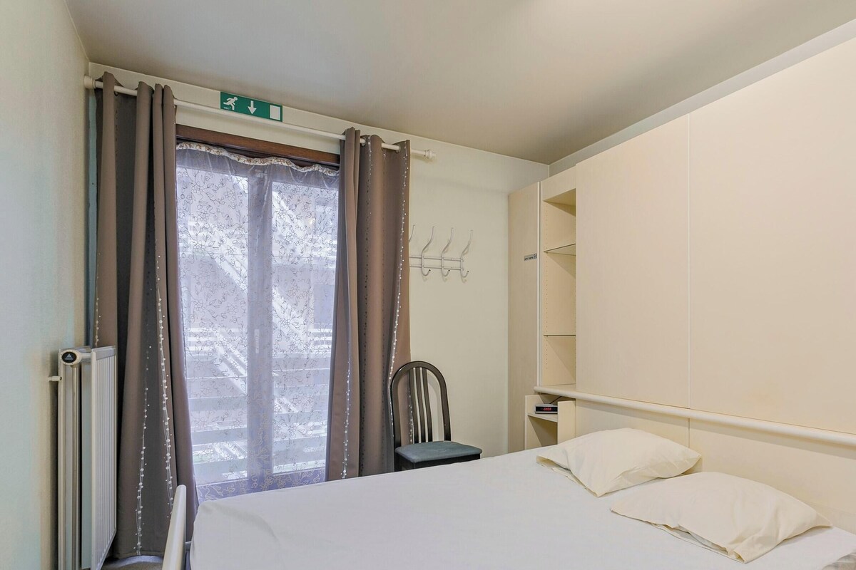 Ein Schlafzimmer mit einem Bett, einem Sessel, einem Heizkörper, einem Fenster mit Vorhängen und einem Regal.