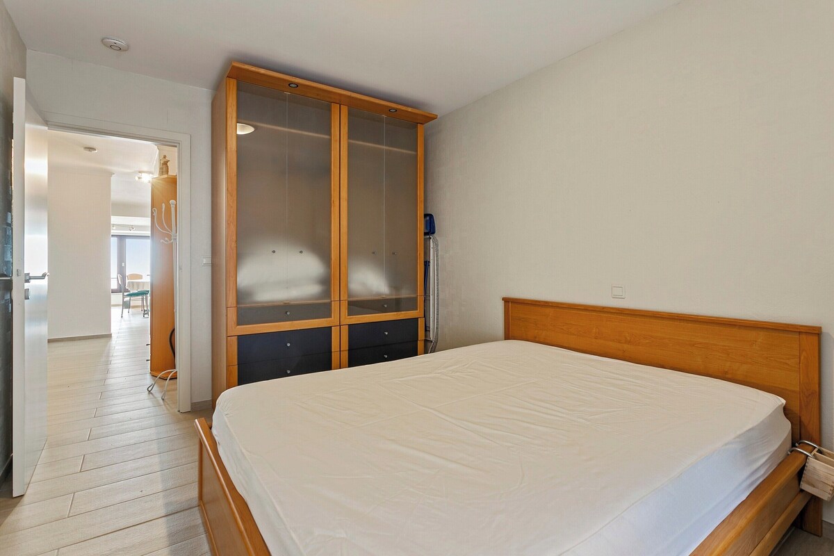 Ein Schlafzimmer mit Holzbettgestell, einem großen Kleiderschrank mit Glastür und einem sichtbaren Badezimmer im Hintergrund.