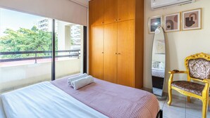 Un dormitorio con cama, armario, silla y balcón con vistas a edificios y árboles.
