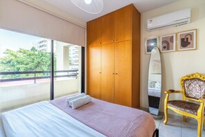 Ein Schlafzimmer mit Bett, Schrank, Stuhl und einem Balkon mit Blick auf Gebäude und Bäume.