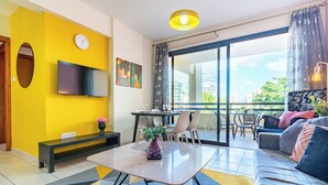 Una sala de estar moderna con un televisor de pantalla plana, un comedor con mesa y sillas, un sofá con cojines, un ventanal con vistas y una pared de acento amarilla.