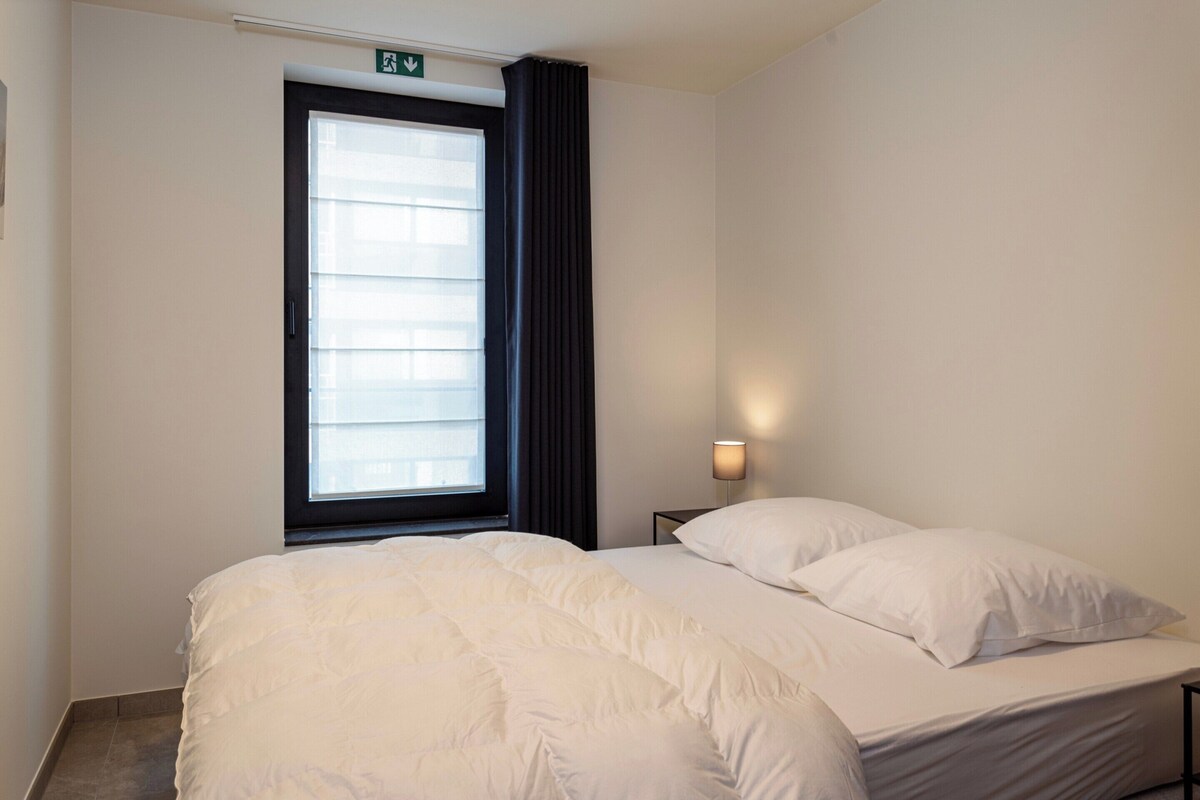 Ein Hotelzimmer mit Bett, Fenster mit Jalousien, einer Wandleuchte und einer Tür mit Ausgangsschild.
