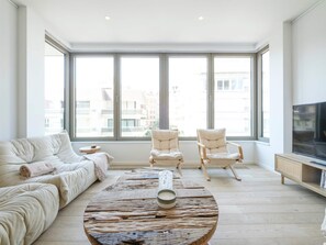 A spacious living room with a large window, a round coffee table, a sofa, two armchairs, and a television. - Apartment Knokke-heist With Sea View (Knokke-Heist)