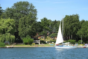 Ein Segelboot auf einem See, im Hintergrund ein Park und Gebäude.