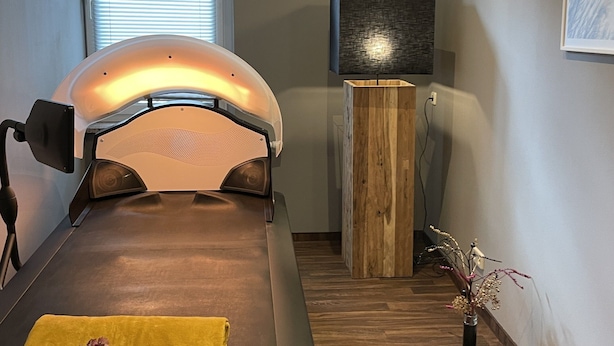 A treatment room with a massage table, a wooden cabinet, a wall-mounted TV, and a window with blinds. - Seepark Heidenholz, Plau am See (Plau am See)