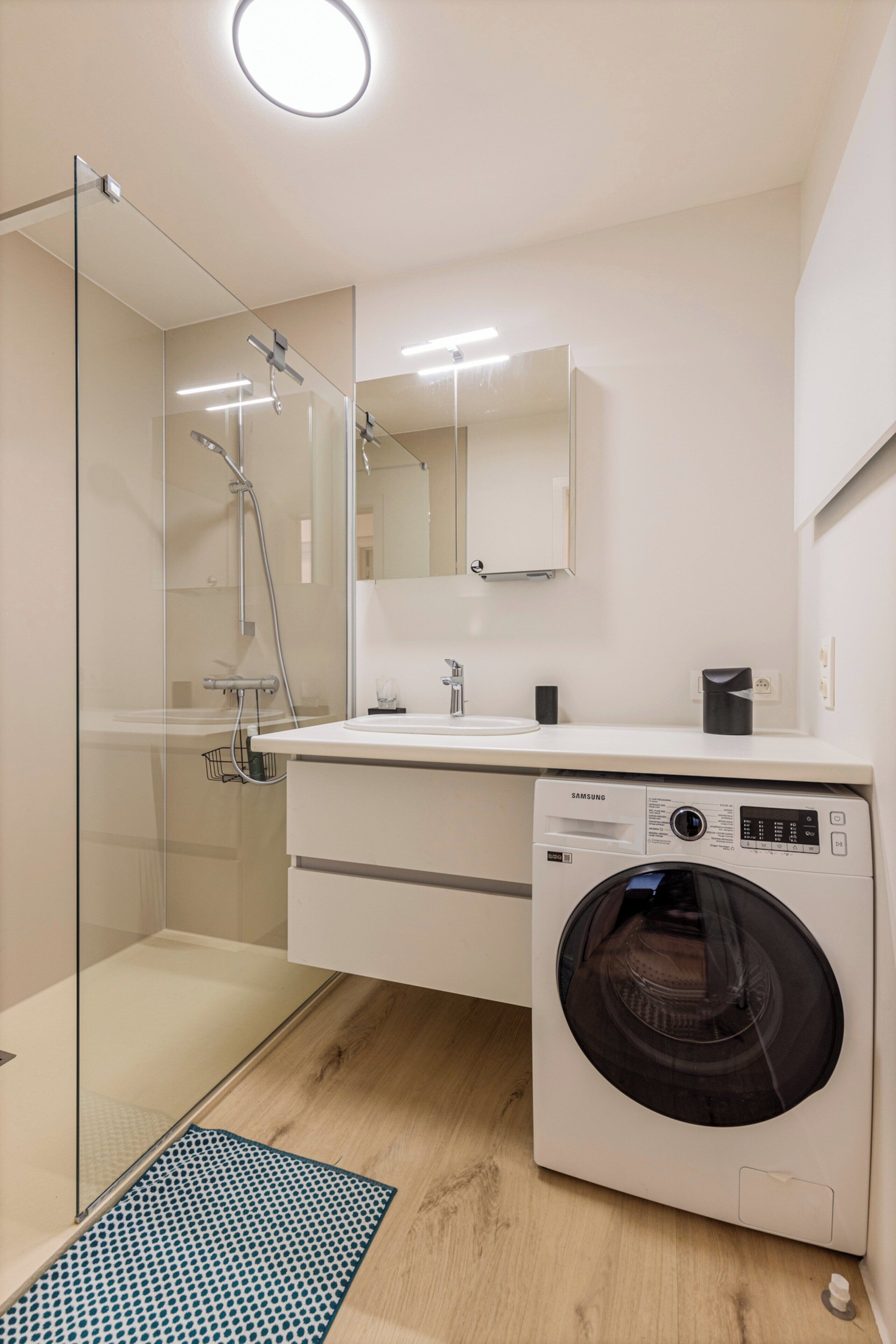 A modern bathroom with a glass shower enclosure, a washing machine, and a white sink with a mirror above.