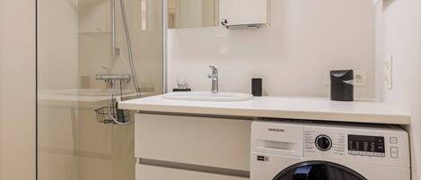 A modern bathroom with a glass shower enclosure, a washing machine, and a white sink with a mirror above.