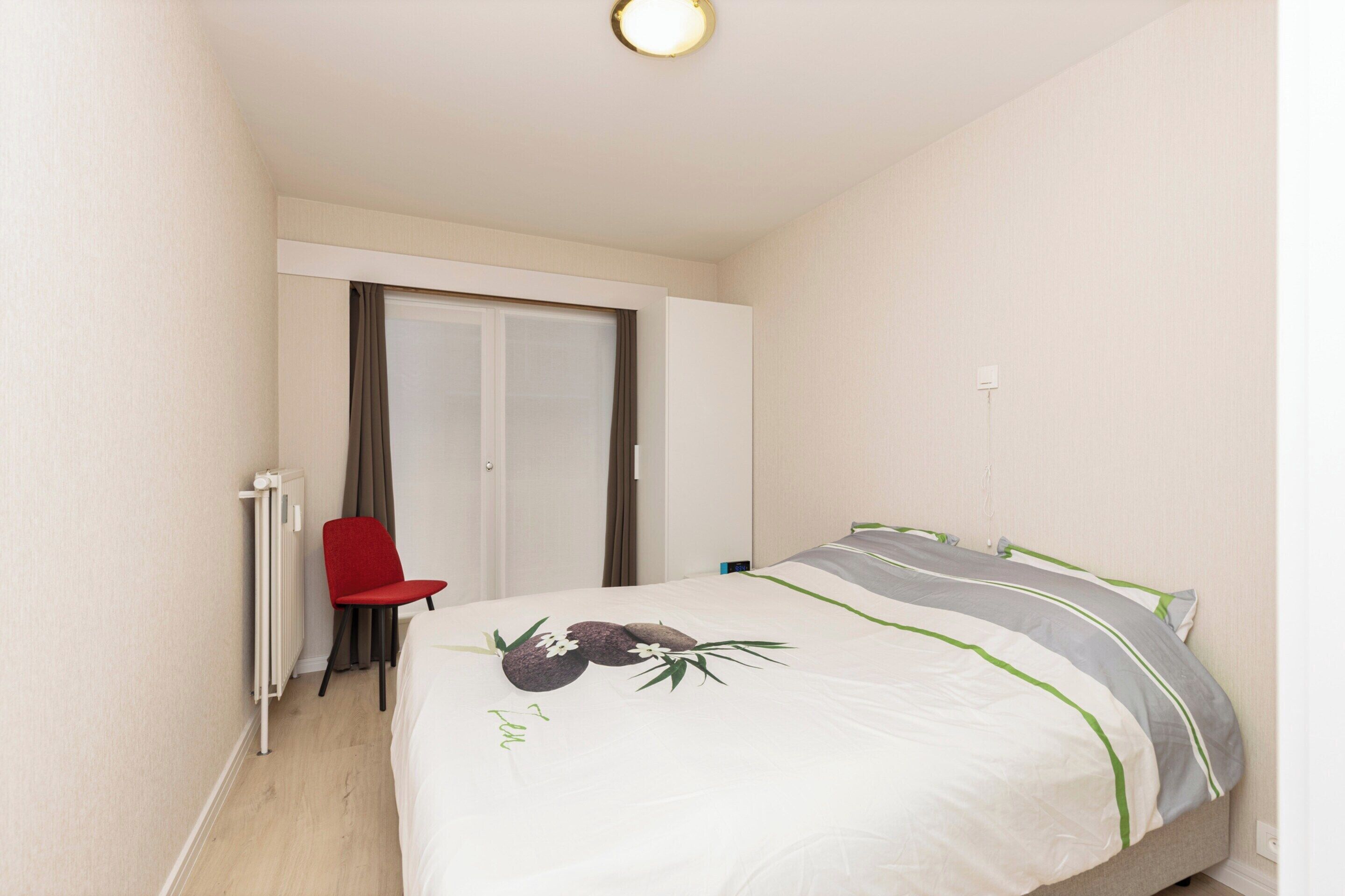 Hotellihuone, jossa on sänky, punainen tuoli, patteri ja ovi.