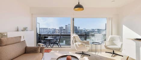 En moderne stue med sofa, to stole, et lille bord og et stort vindue med udsigt over byen.