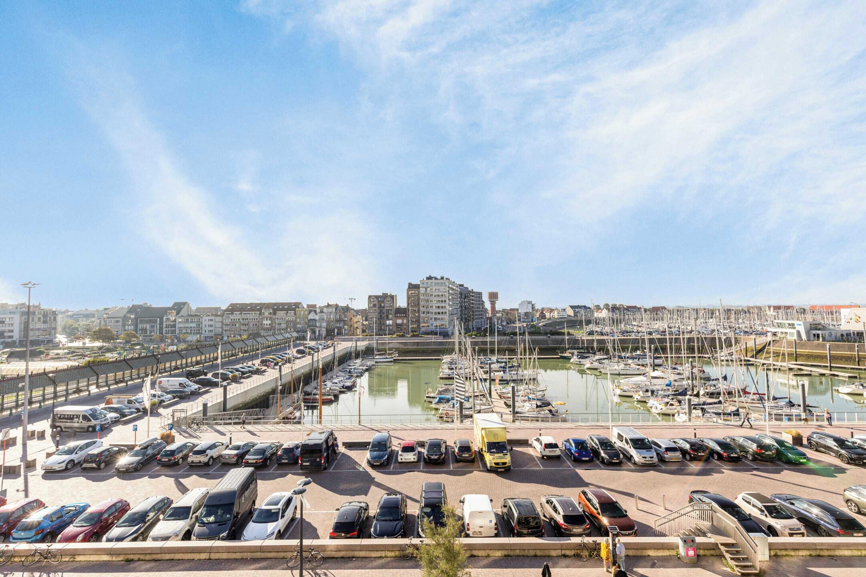 A marina with parked cars and boats, a clear sky, and buildings in the background.