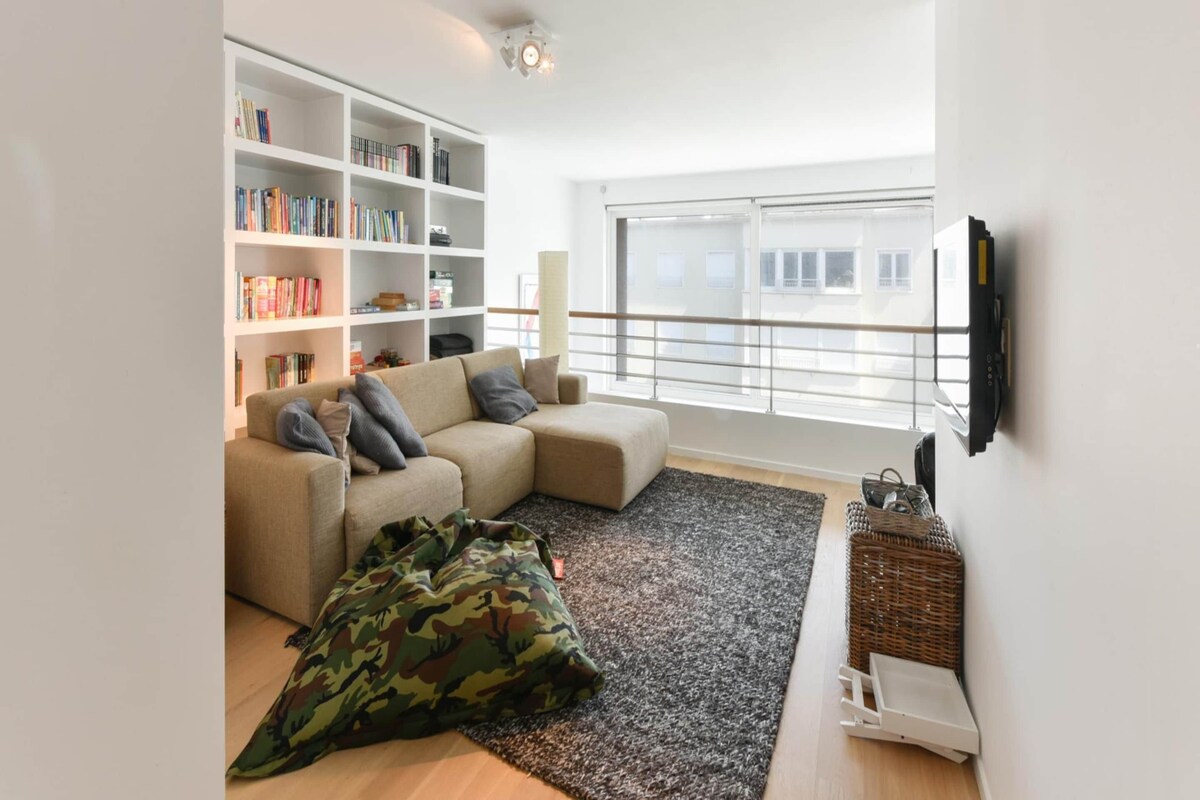 A modern living room with a sectional sofa, a bookshelf, a flat-screen TV, and a large window.