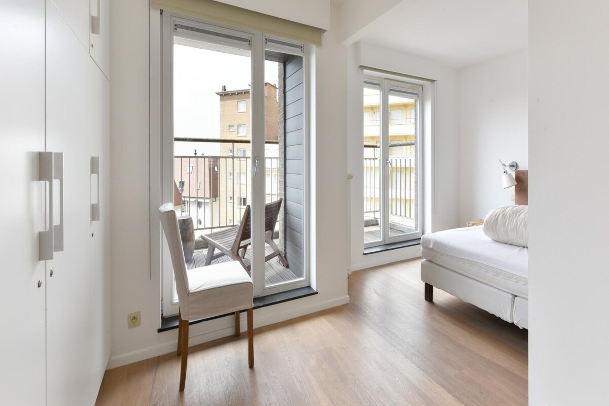 Ein modernes Schlafzimmer mit Bett, Sessel und einem Balkon mit Blick auf Gebäude.