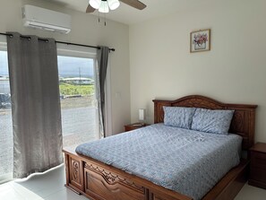 A bedroom with a wooden bed, a nightstand, a wall-mounted air conditioner, and a window with curtains. - Spacious 4-bedroom building in gorgeous Hilo with AC. Center of Hilo. (Hilo)