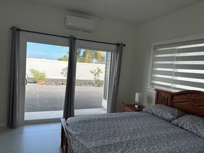 A bedroom with a bed, a nightstand, a window with blinds, and an air conditioning unit. - Spacious 4-bedroom building in gorgeous Hilo with AC. Center of Hilo. (Hilo)