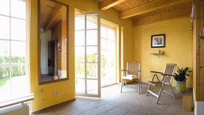 Una habitación con ventanas amplias, techo de madera, una silla, una planta en maceta y un cuadro en la pared.