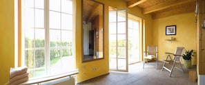 A room with large windows, a wooden ceiling, a chair, a potted plant, and a framed picture on the wall. - Hof Kracht – "Jaromar" One-Bedroom Apartment for 2 People on the Baltic Sea (Putgarten)