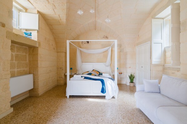 A bedroom with a four-poster bed, a white sofa, a potted plant, and a stone wall.