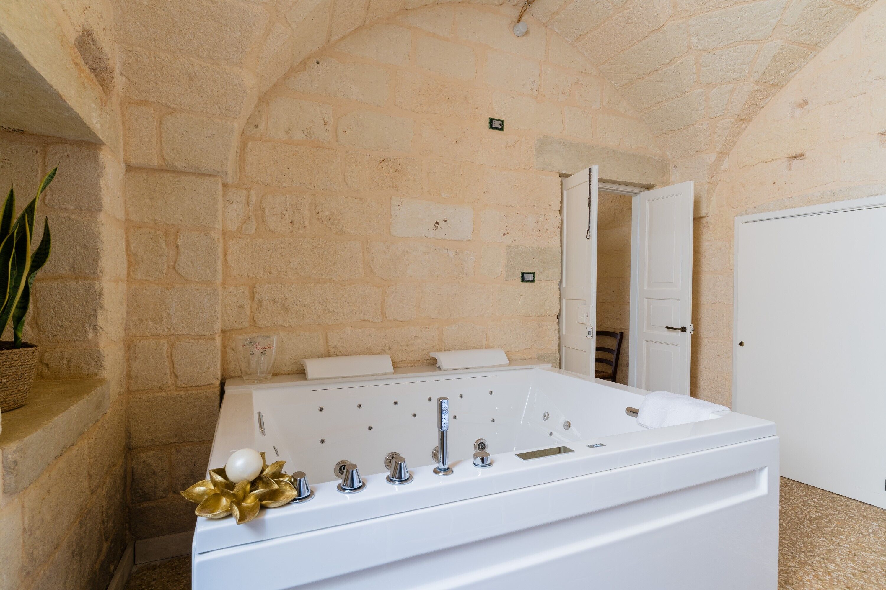 A spa room with a white bathtub, stone walls, a potted plant, and a door leading to another room.