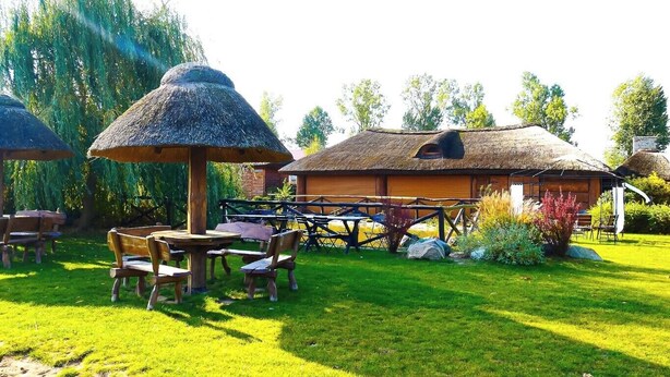 Un espacio de estar al aire libre de estilo rústico con mesas y sillas de madera, rodeado de sombrillas de paja, y un edificio de madera con techo de paja.
