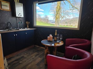 Une cuisine avec une fenêtre donnant sur l’extérieur, un évier, des placards, une table avec des bougies et des verres, et une chaise rouge.