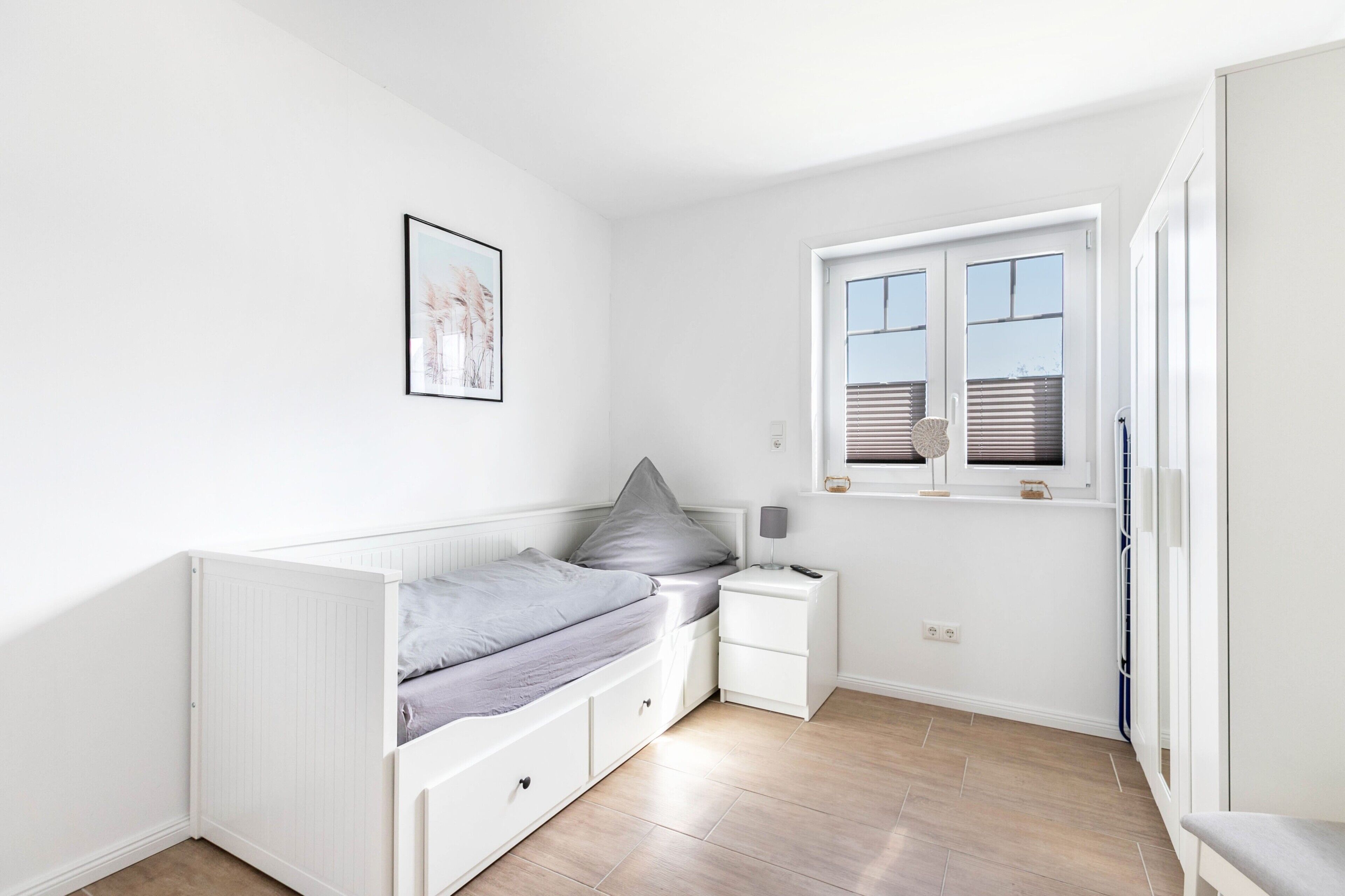A small, bright room with a single bed, a nightstand, a window with blinds, and a framed picture on the wall.