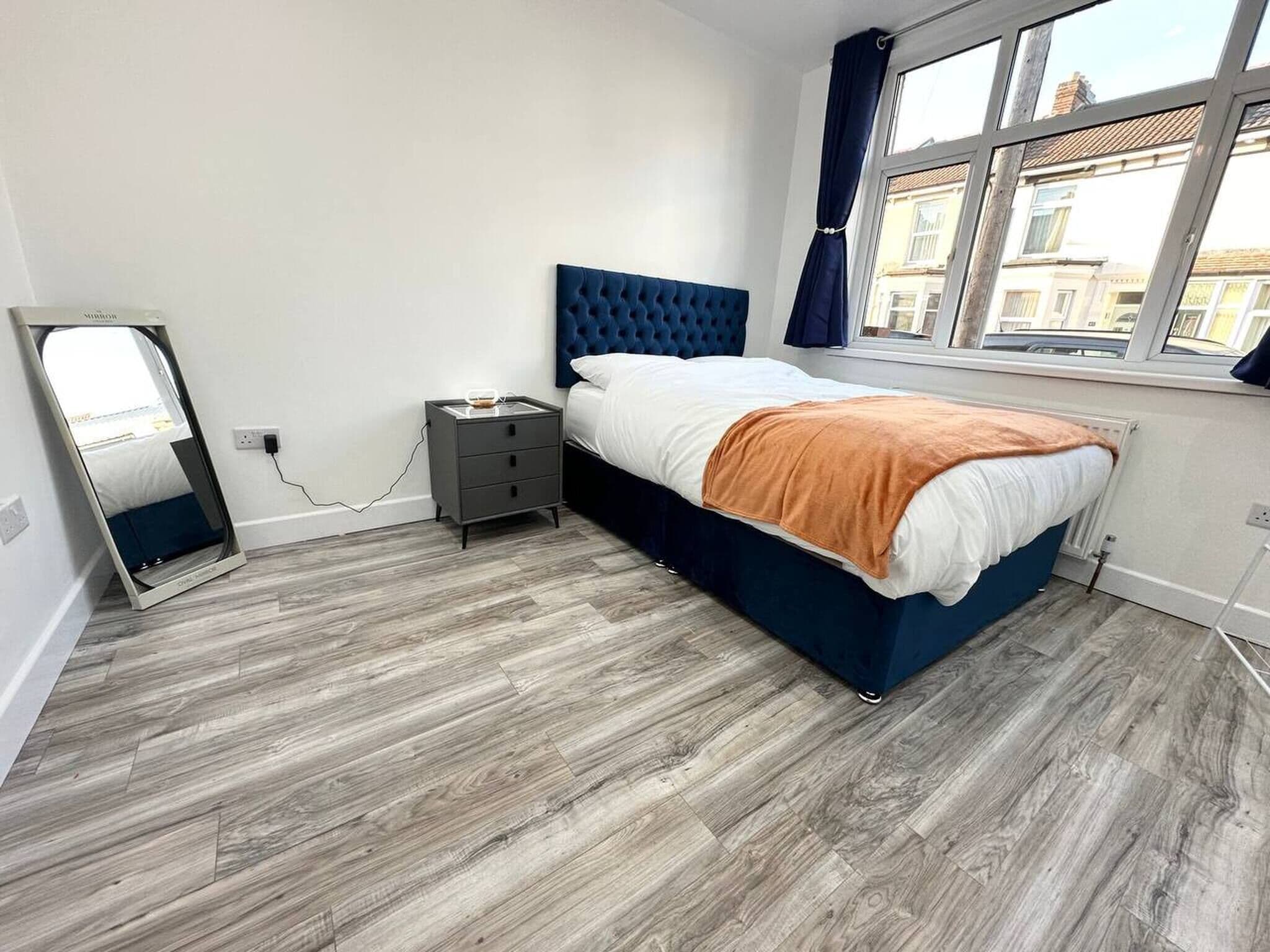 A bedroom with a bed, a bedside table, a mirror, and a window with curtains.