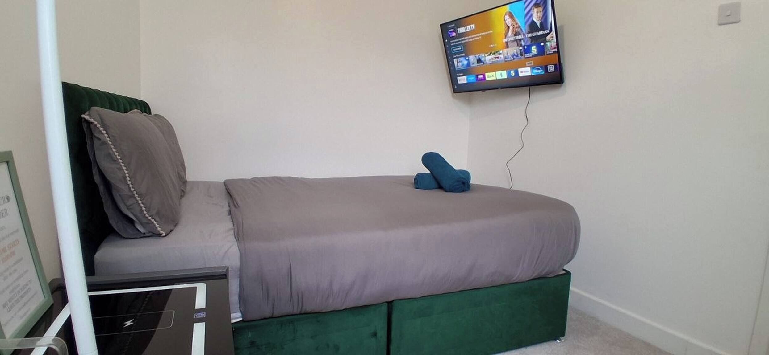 A single bed with a green upholstered headboard, a flat-screen TV mounted on the wall, and a power outlet on the wall.