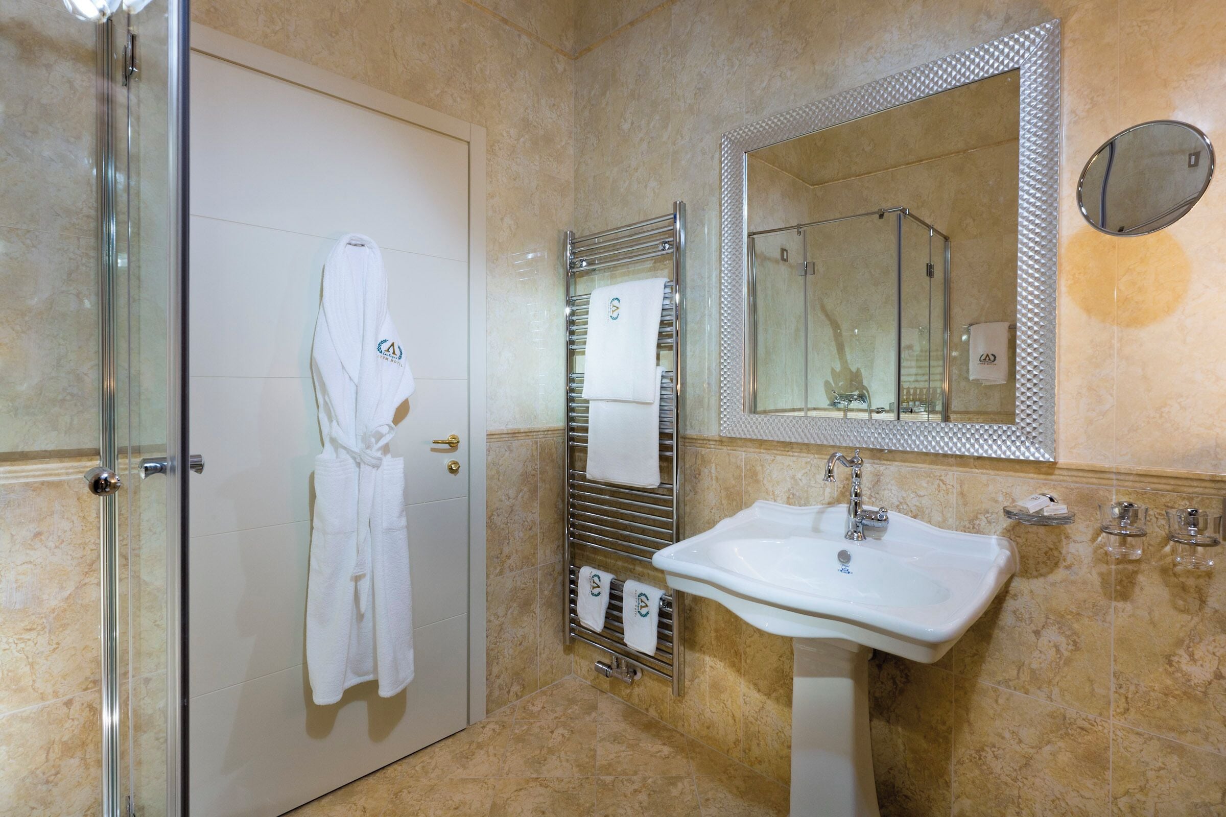Un baño con cabina de ducha de vidrio, un lavabo blanco, un perchero con toallas y un espejo grande.