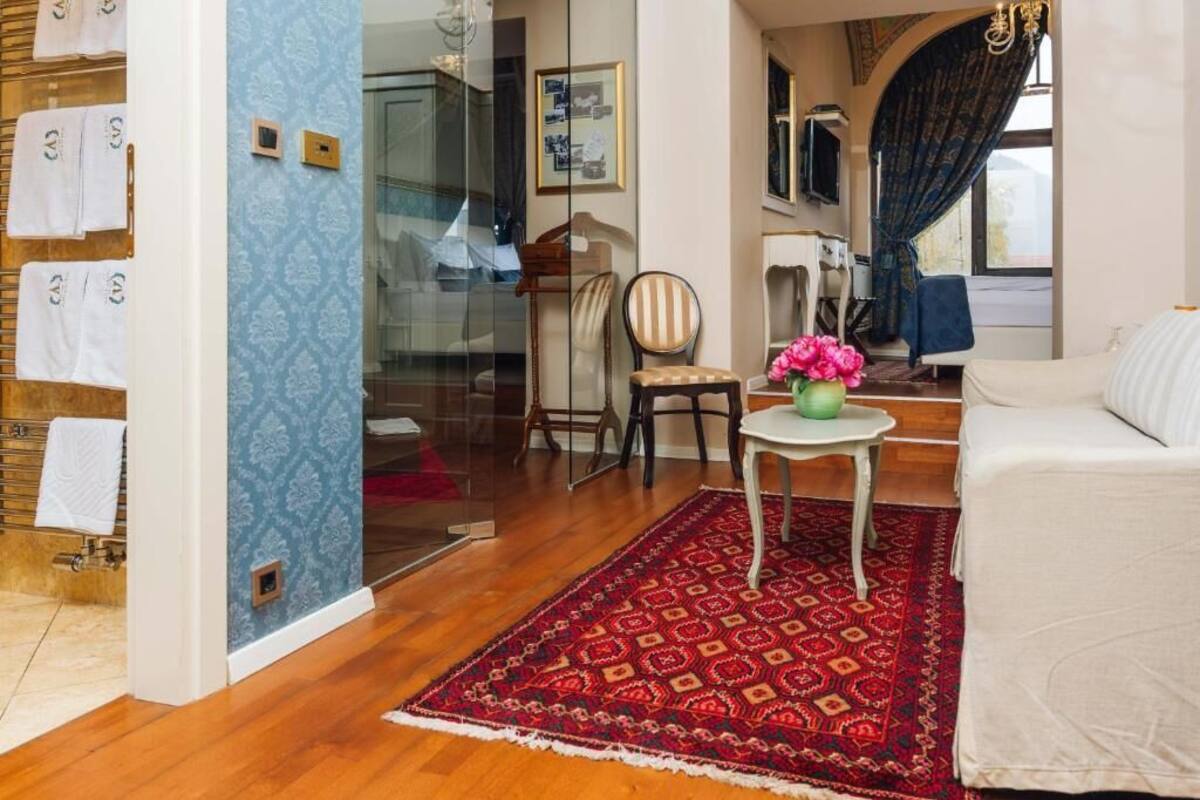 A hotel room with a wooden floor, a red patterned rug, a white sofa, a small table with a vase of flowers, and a chair.