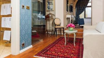 A hotel room with a wooden floor, a red patterned rug, a white sofa, a small table with a vase of flowers, and a chair.