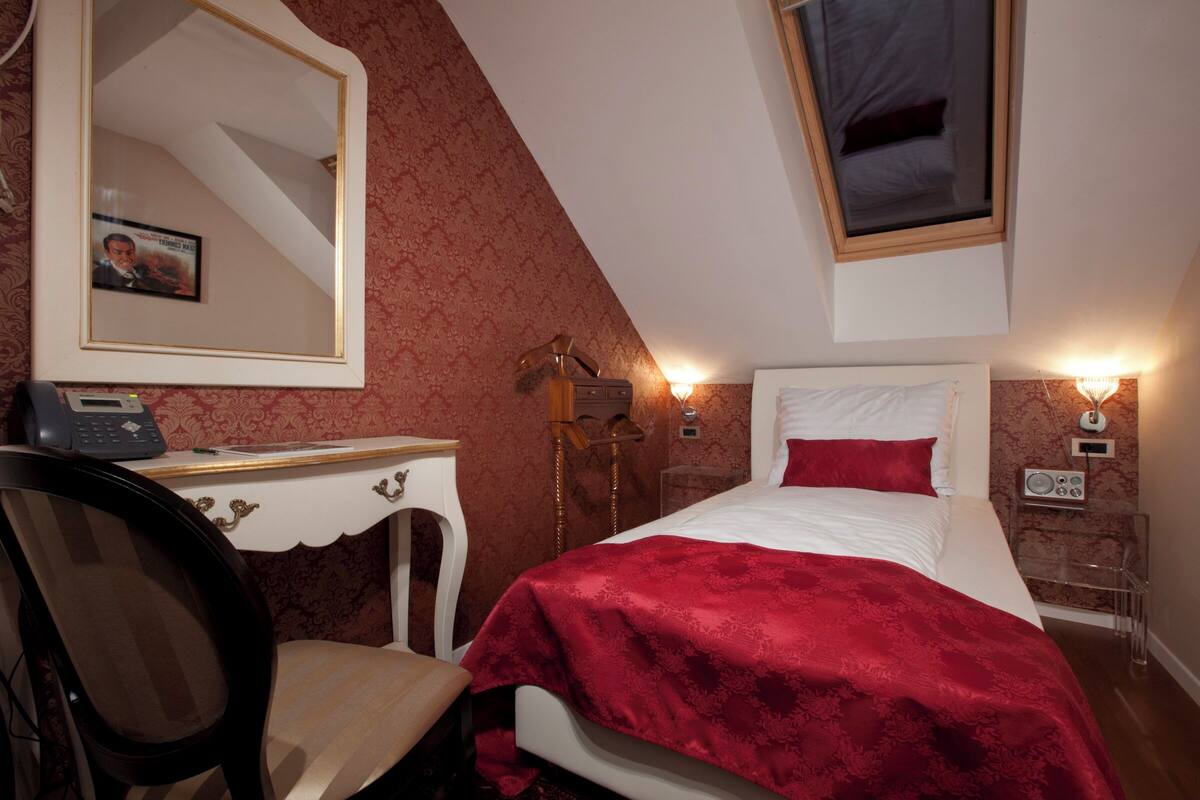 A bedroom with a bed, a desk, a chair, a mirror, a bedside table, a lamp, and a radio.