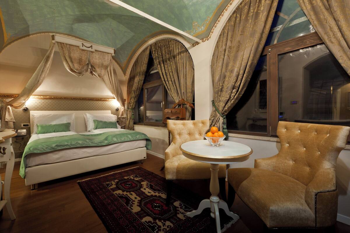 A bedroom with a four-poster bed, a round table with a bowl of fruit, and a tufted armchair.