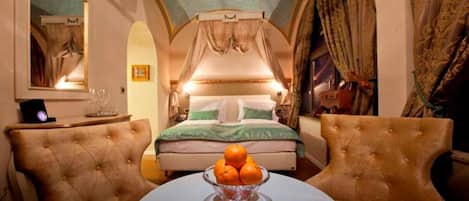 A bedroom with a four-poster bed, a round table with a bowl of fruit, and two armchairs.
