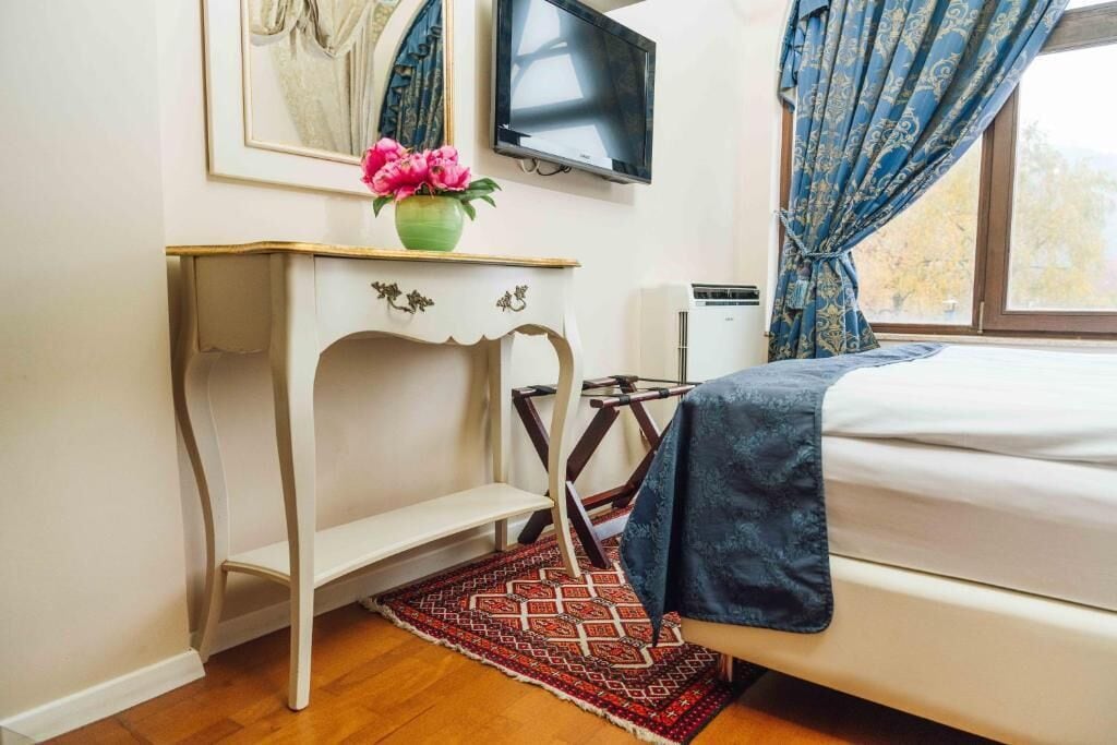 A bedroom with a bed, a TV mounted on the wall, a small table with a vase of flowers, and a window with curtains.