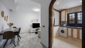 A modern kitchen with a washing machine, a dining area with a table and chairs, and a TV in the background. - Vacation Apartment 9 min from Bajo de Guía with Shared Terrace, Wi-Fi & Air Conditioning (Sanlúcar de Barrameda)