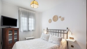 A bedroom with a bed, a television on a cabinet, a bedside table with a lamp, and decorative wall art. - Vacation Apartment 9 min from Bajo de Guía with Shared Terrace, Wi-Fi & Air Conditioning (Sanlúcar de Barrameda)