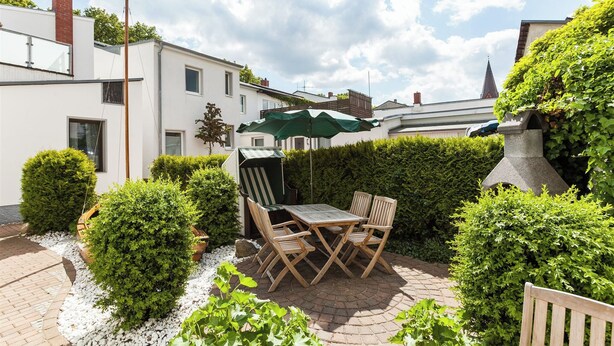 Eine Terrasse mit Tisch und Stühlen, umgeben von Hecken und einem Pflasterweg.