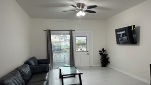 A living room with a black leather sofa, a glass coffee table, a ceiling fan, a TV mounted on the wall, and a sliding glass door leading to an outdoor area. - Spacious 4-bedroom , newly building in center of Hilo. (Hilo)