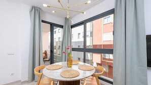 Et moderne spiseområde med et rundt bord, træstole, en vase med blomster og et stort vindue med gardiner.