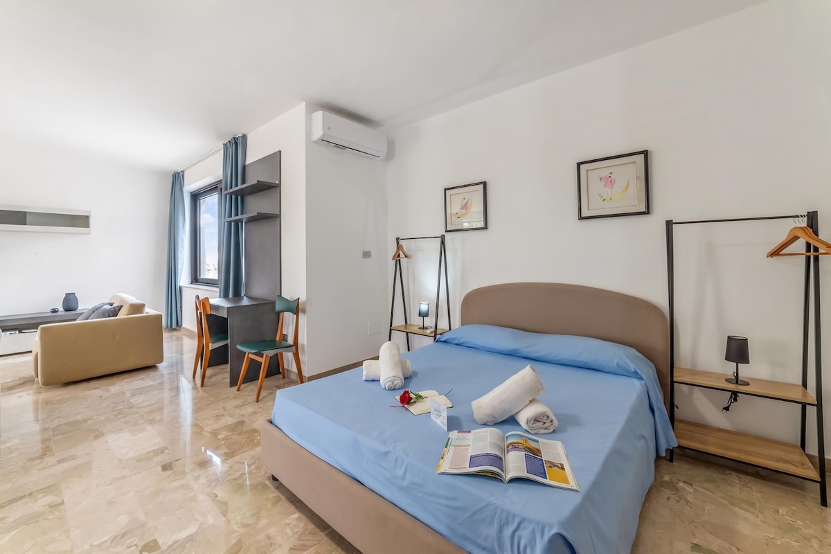 A modern hotel room with a bed, a desk, a chair, a wardrobe, and a bedside table.