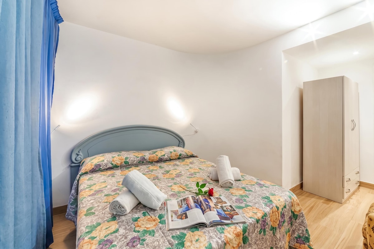 A neatly made bed with a floral bedspread, two rolled towels, and a magazine on the bed.