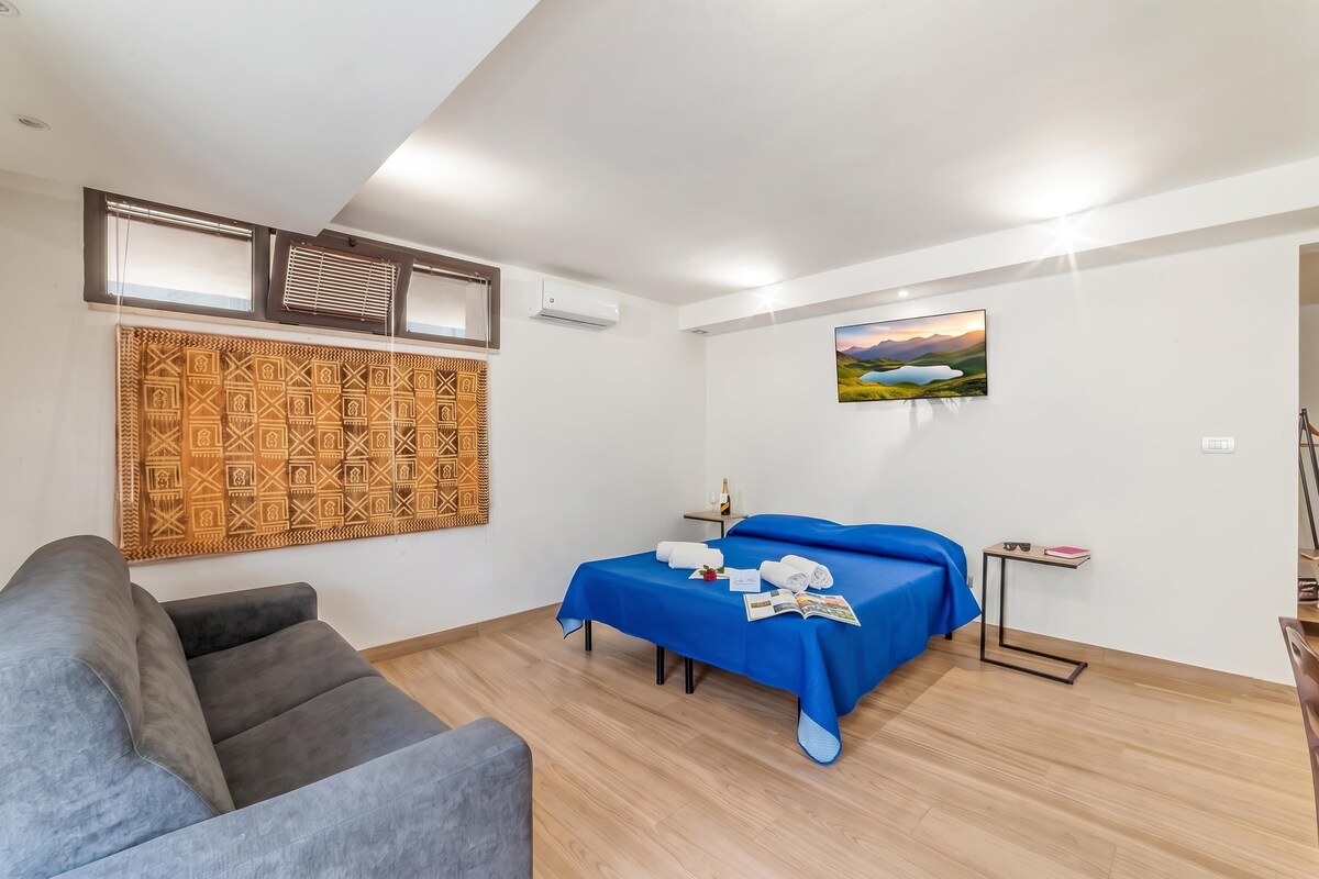 A room with a bed, a sofa, a small table with a bottle and glasses, and a wall-mounted air conditioner.