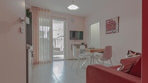 Een moderne woonkamer met een rode bank, een eethoek met witte stoelen, een aan de muur bevestigde televisie en een schuifpui naar een balkon.