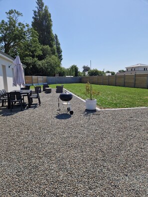 A patio area with a barbecue grill, outdoor seating, and a white building in the background. - Villa Sansouci (Gouville-sur-Mer)